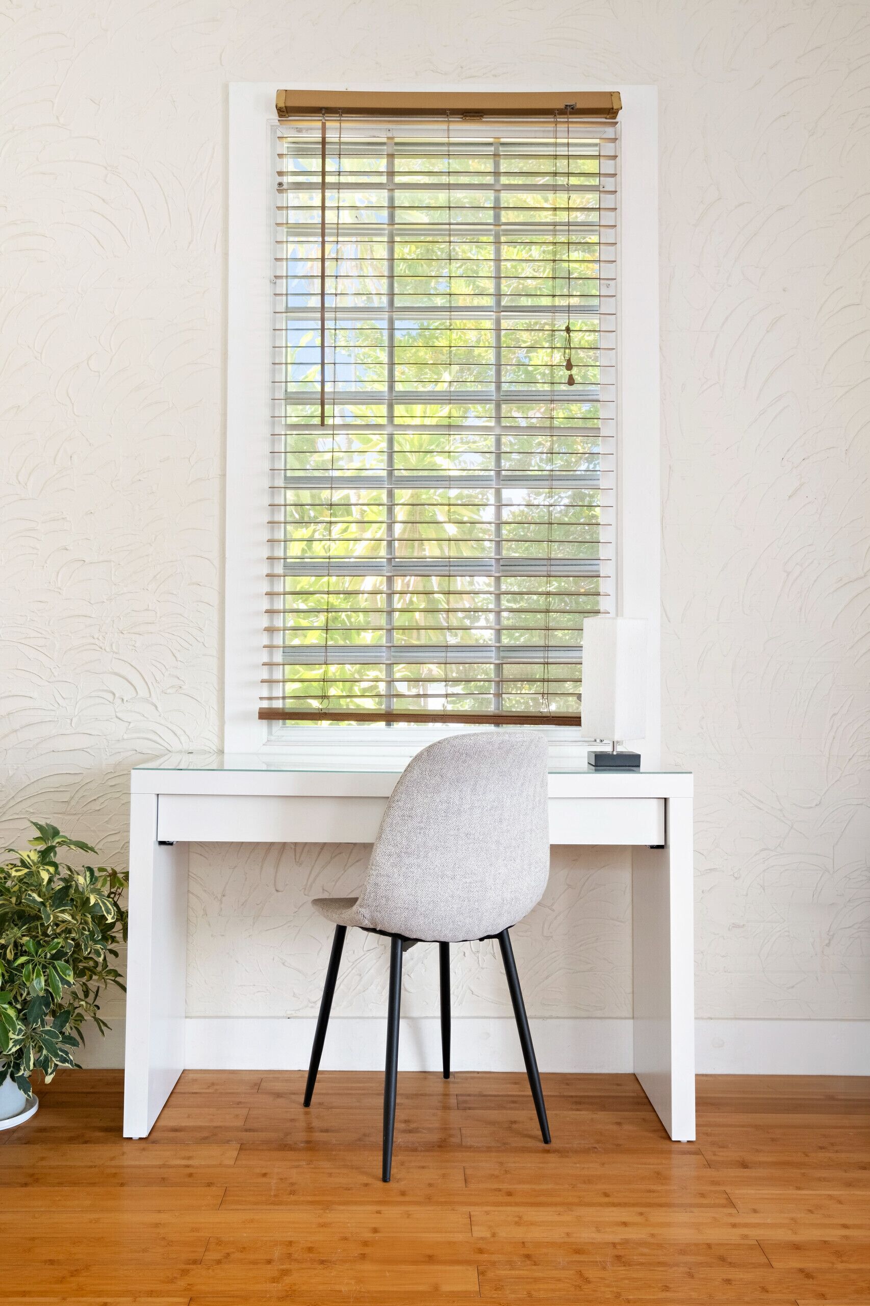 A white desk with a grey chair, a window with blinds, and a potted plant.