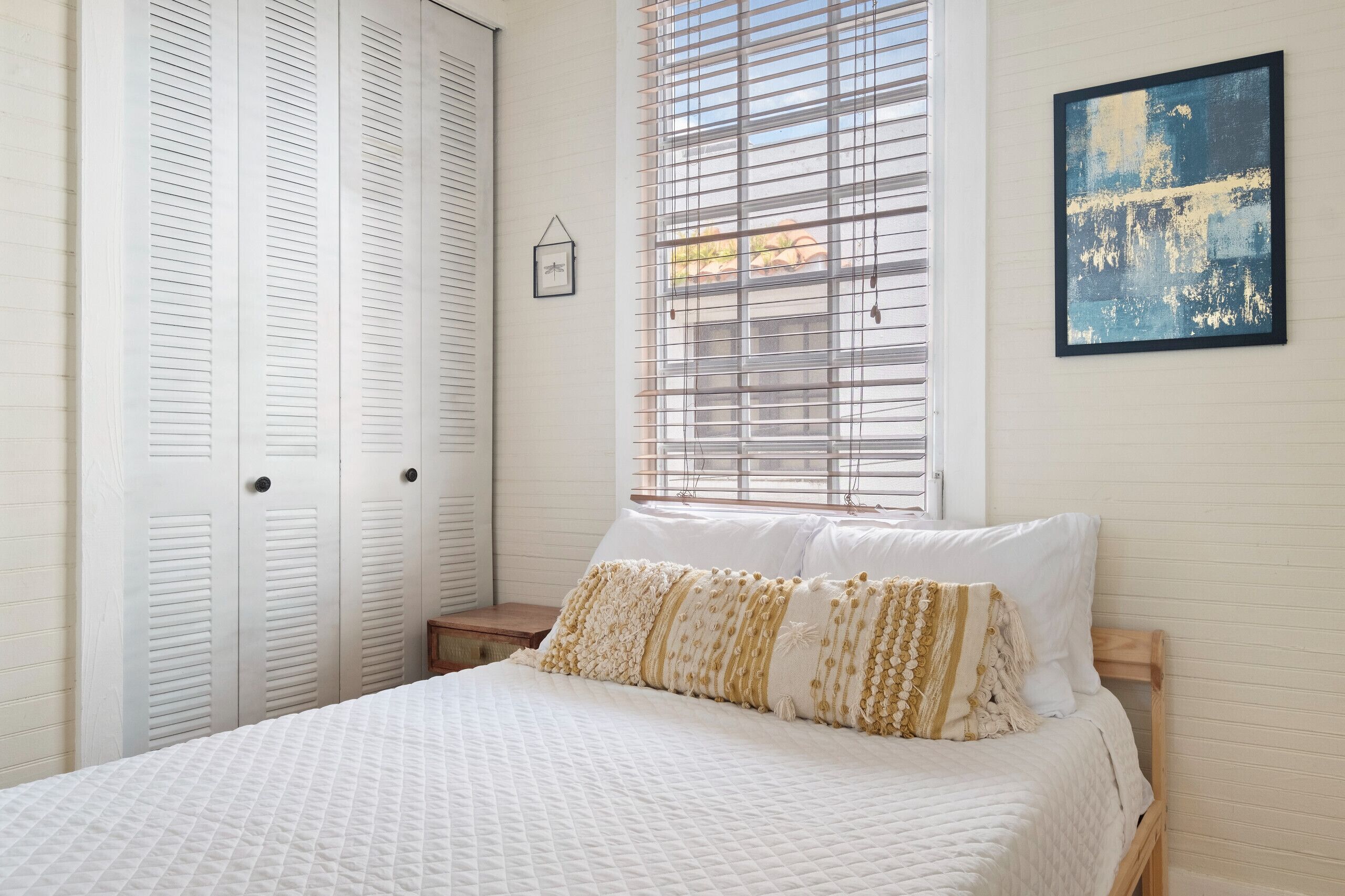 A bedroom with a bed, white walls, a window with blinds, a framed picture, and a closet with sliding doors.