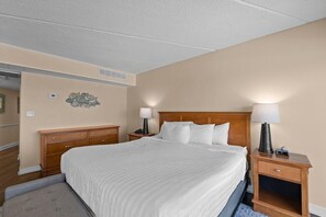 A hotel room with a large bed, two bedside tables with lamps, a wooden dresser, and a wall-mounted air vent. - Deluxe Oceanfront King Room 3rd Floor (Pawleys Island)