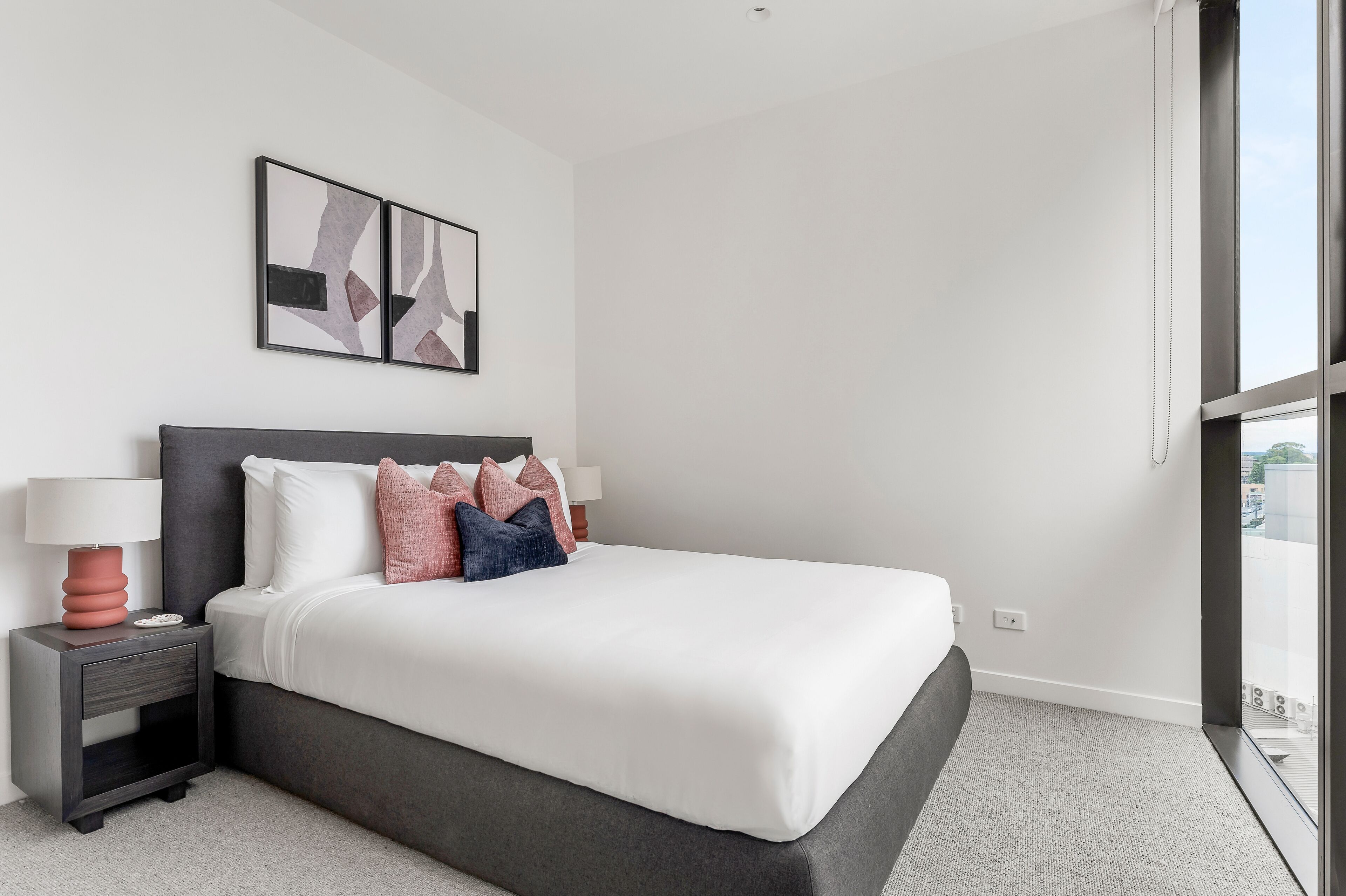 A modern hotel room with a large bed, a nightstand with a lamp, and abstract wall art.