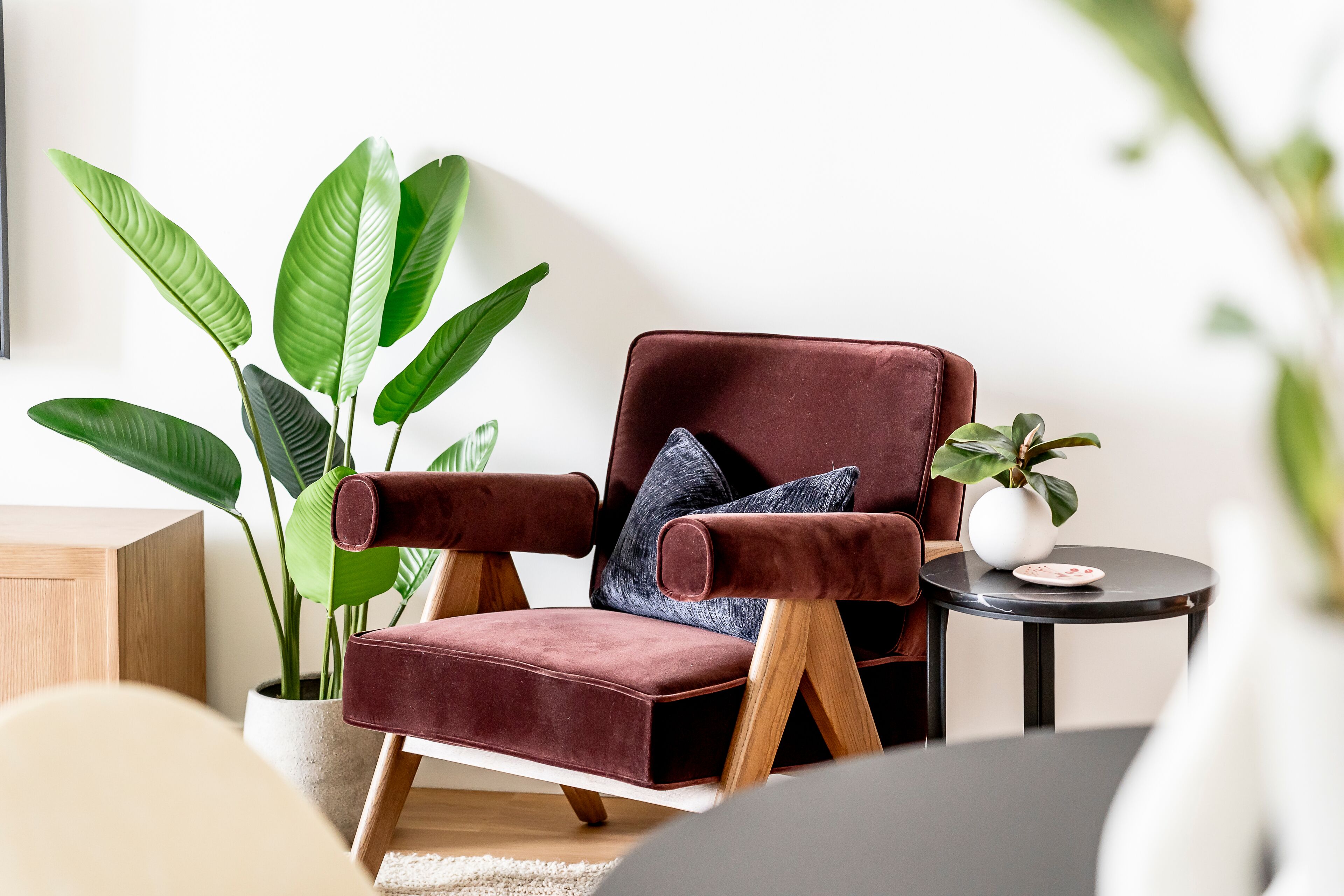 Uma sala de estar moderna com uma poltrona confortável de veludo vermelho, uma grande planta verde e uma pequena mesa redonda com um vaso.