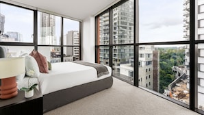 A modern bedroom with a large bed, a nightstand with a lamp, and a view of the city through large windows. - Urban Rest Parramatta Apartments (Harris Park)