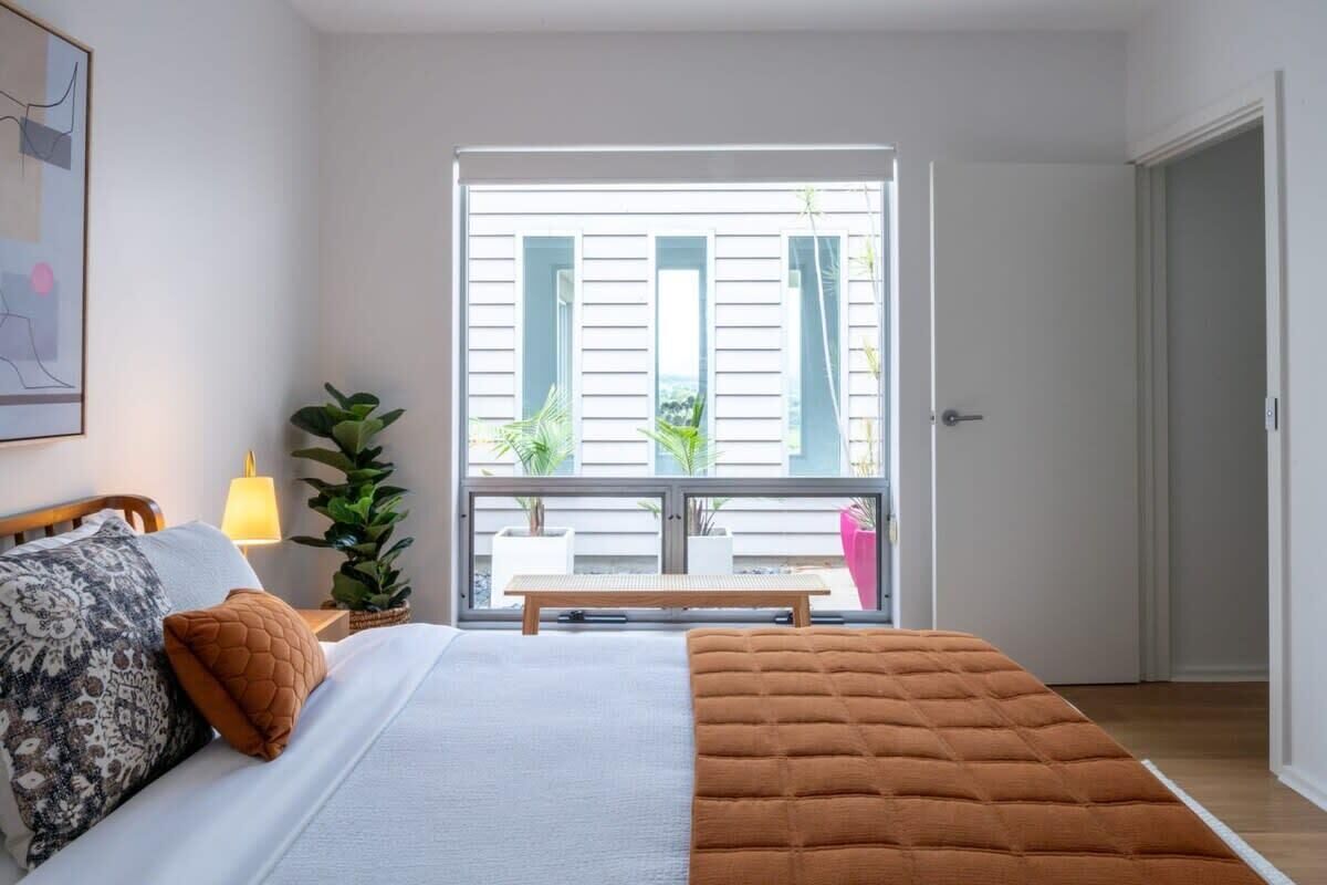 A bedroom with a bed, a bedside table with a lamp, a window with blinds, and a view of the outdoors.