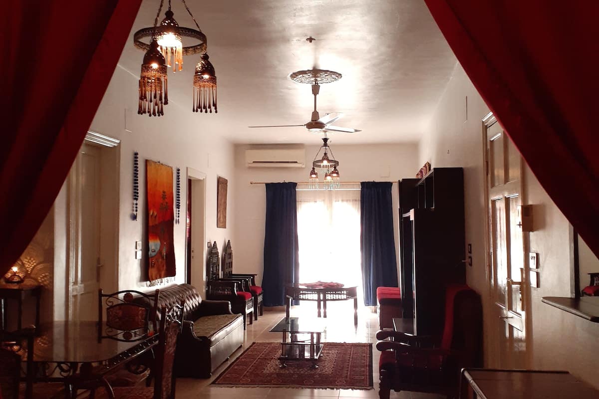 A spacious living room with a high ceiling, a chandelier, and a ceiling fan. There is a dining area with a table and chairs, and a sofa.