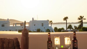 A table with bottles and candles, overlooking a coastal area with white buildings and palm trees. - Anapnoe Resort by ParosKite (Paros)