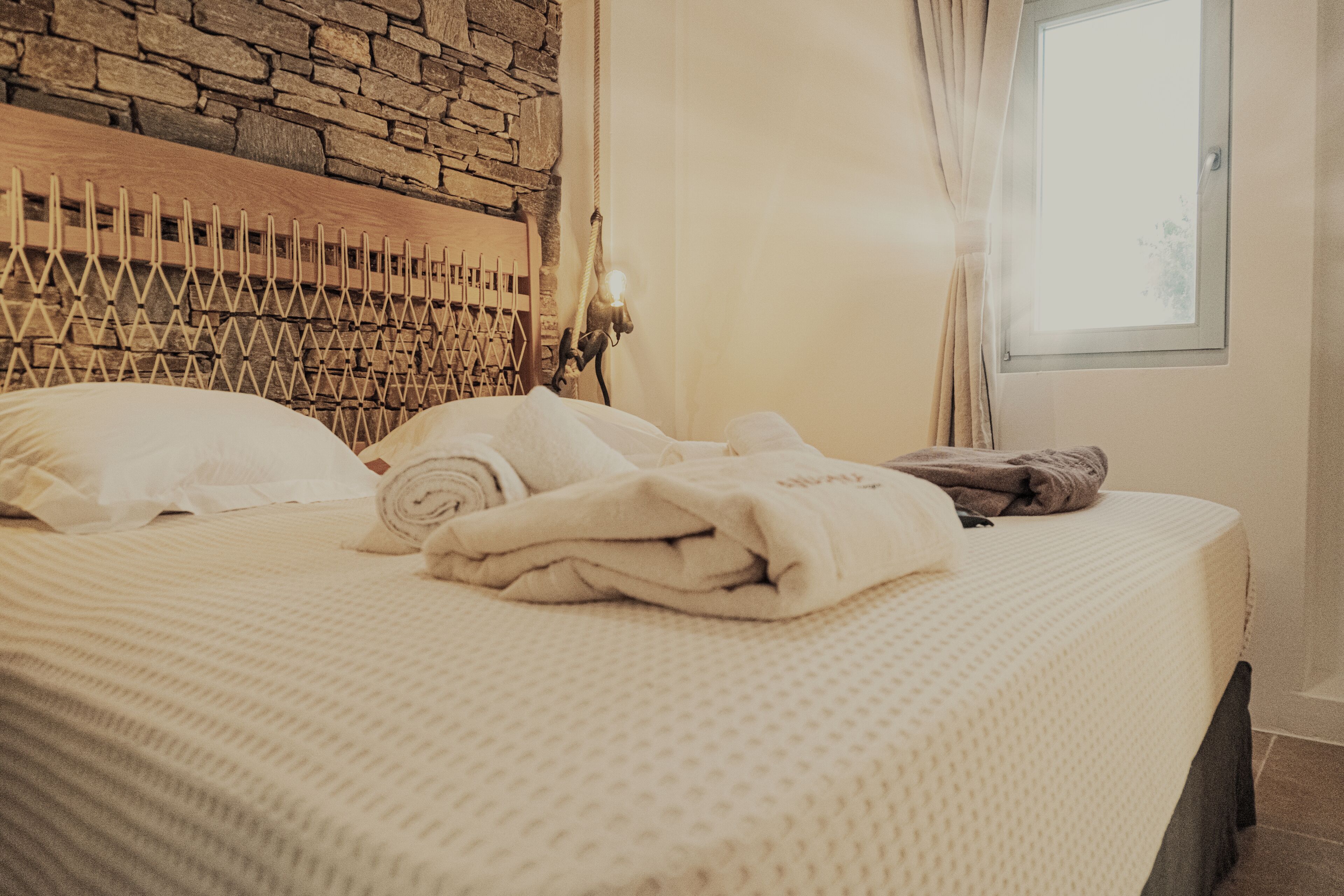 Un letto rifatto con cura, una finestra con tende e una parete in pietra alle spalle del letto.