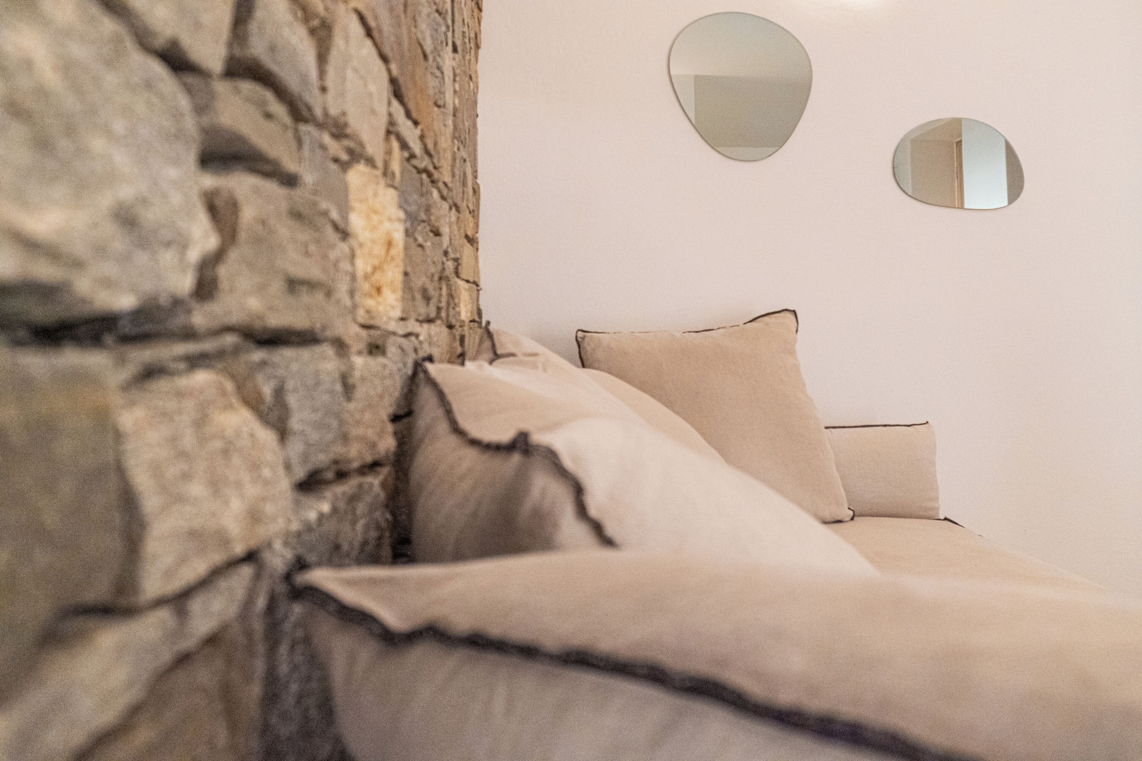 A bed with beige bedding against a stone wall, two wall-mounted mirrors, and a plain white wall.