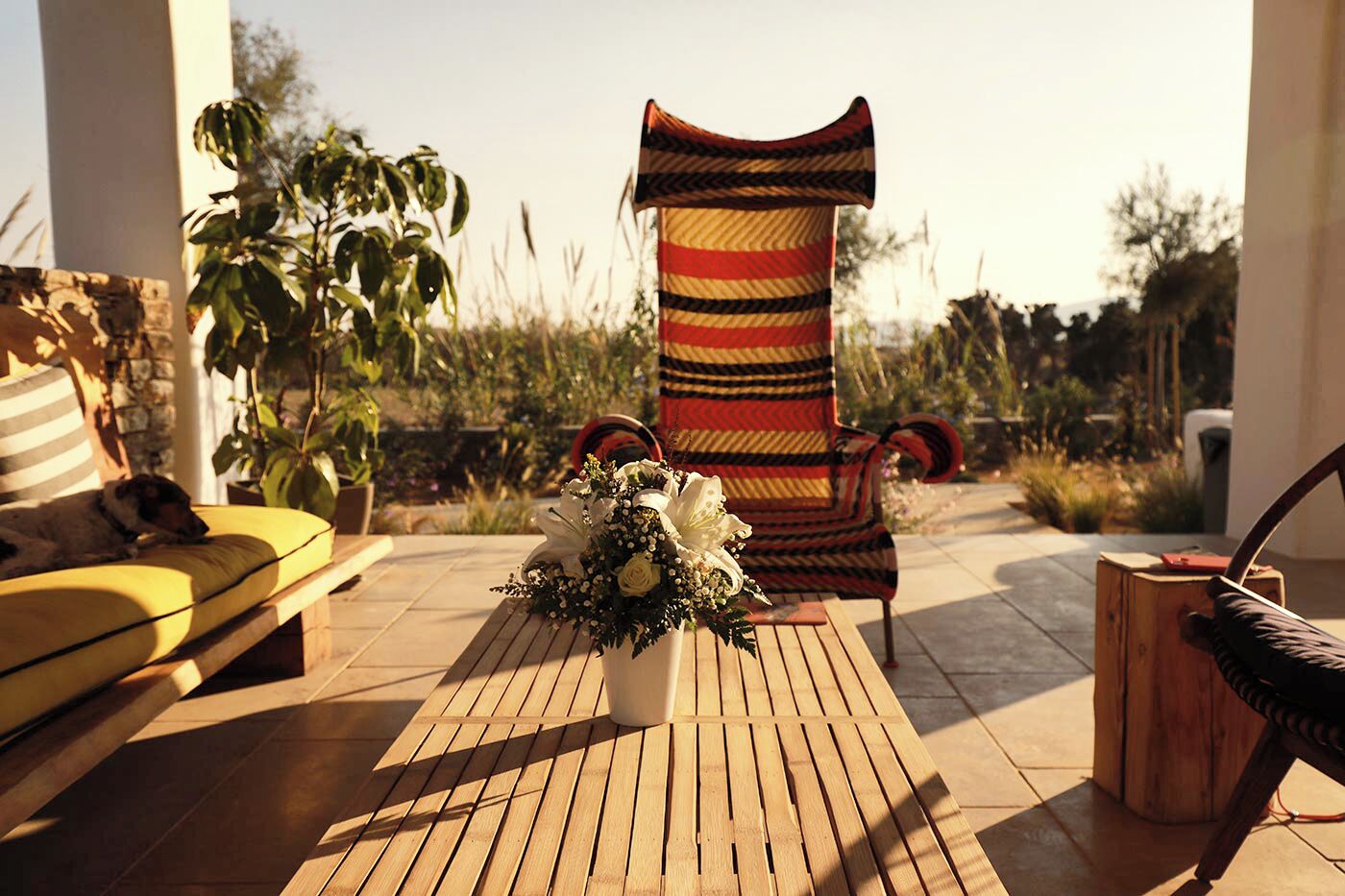 Una terraza de madera con una silla a rayas, un sofá amarillo y una mesa de madera con un arreglo floral.