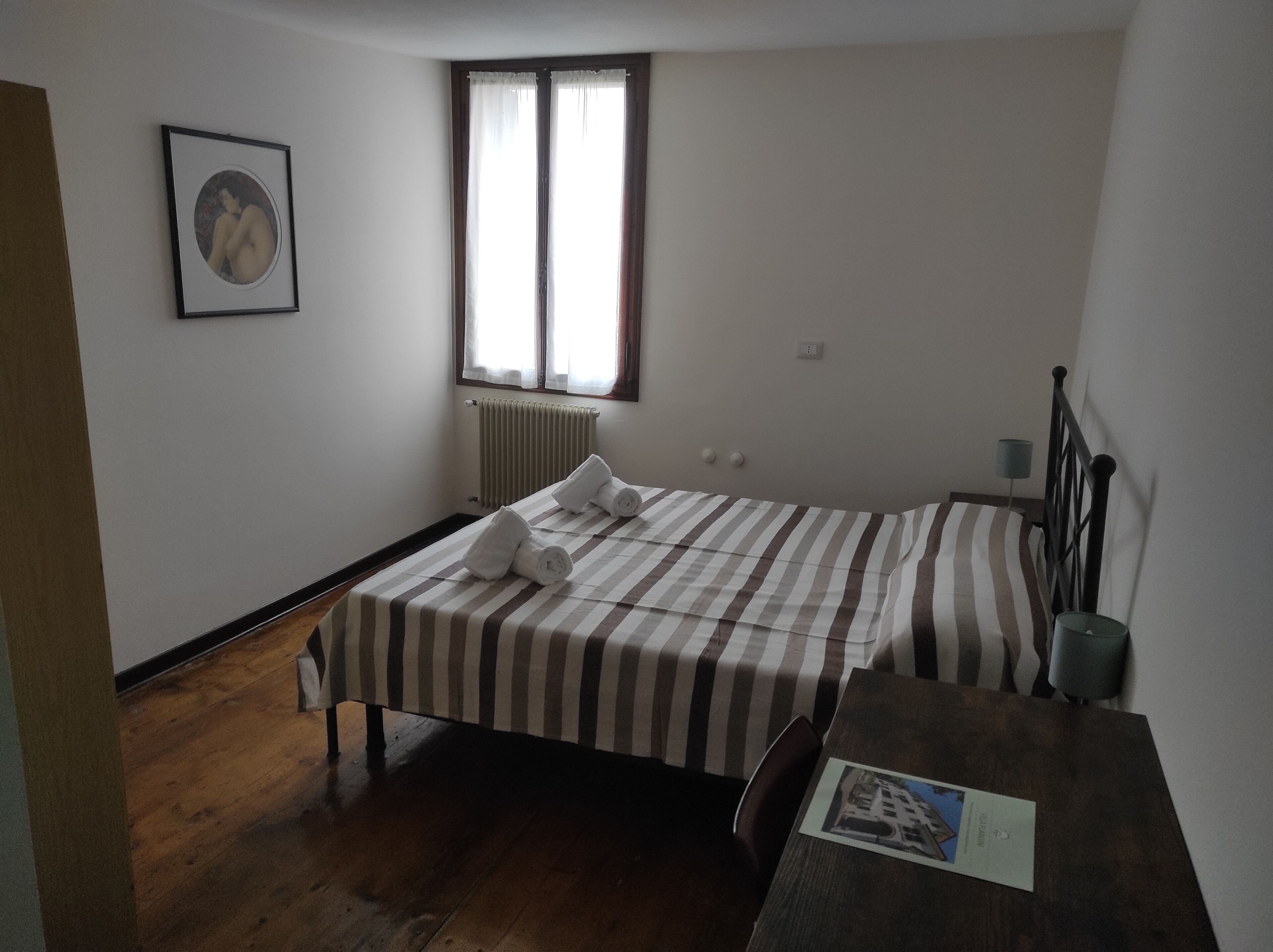 Une petite chambre à coucher, bien rangée, avec un lit simple, un plancher en bois, un radiateur, une fenêtre avec des rideaux et un tableau encadré au mur.