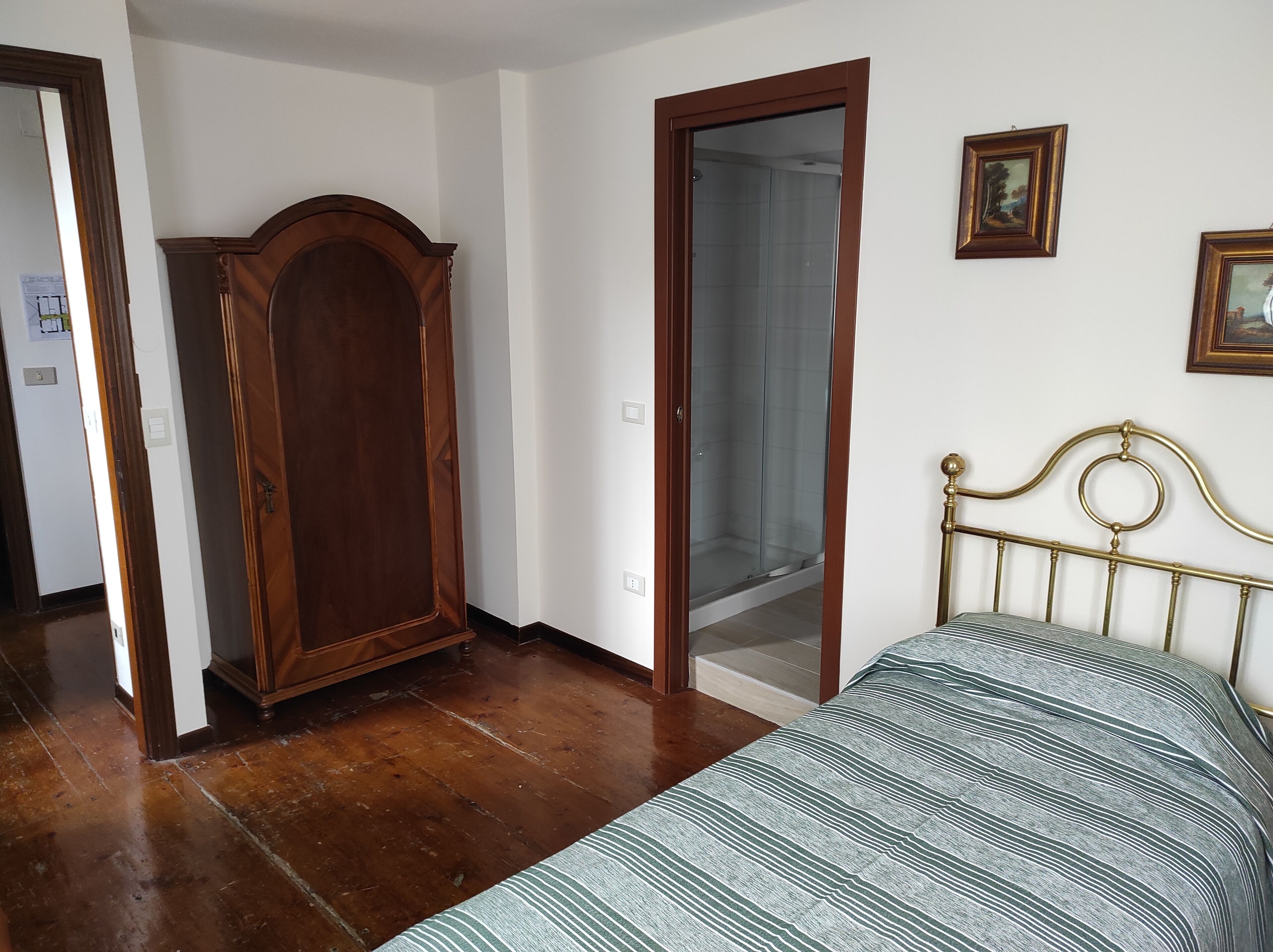 Une chambre avec une armoire en bois, une porte de douche en verre, un lit avec une tête de lit en métal et deux tableaux encadrés au mur.