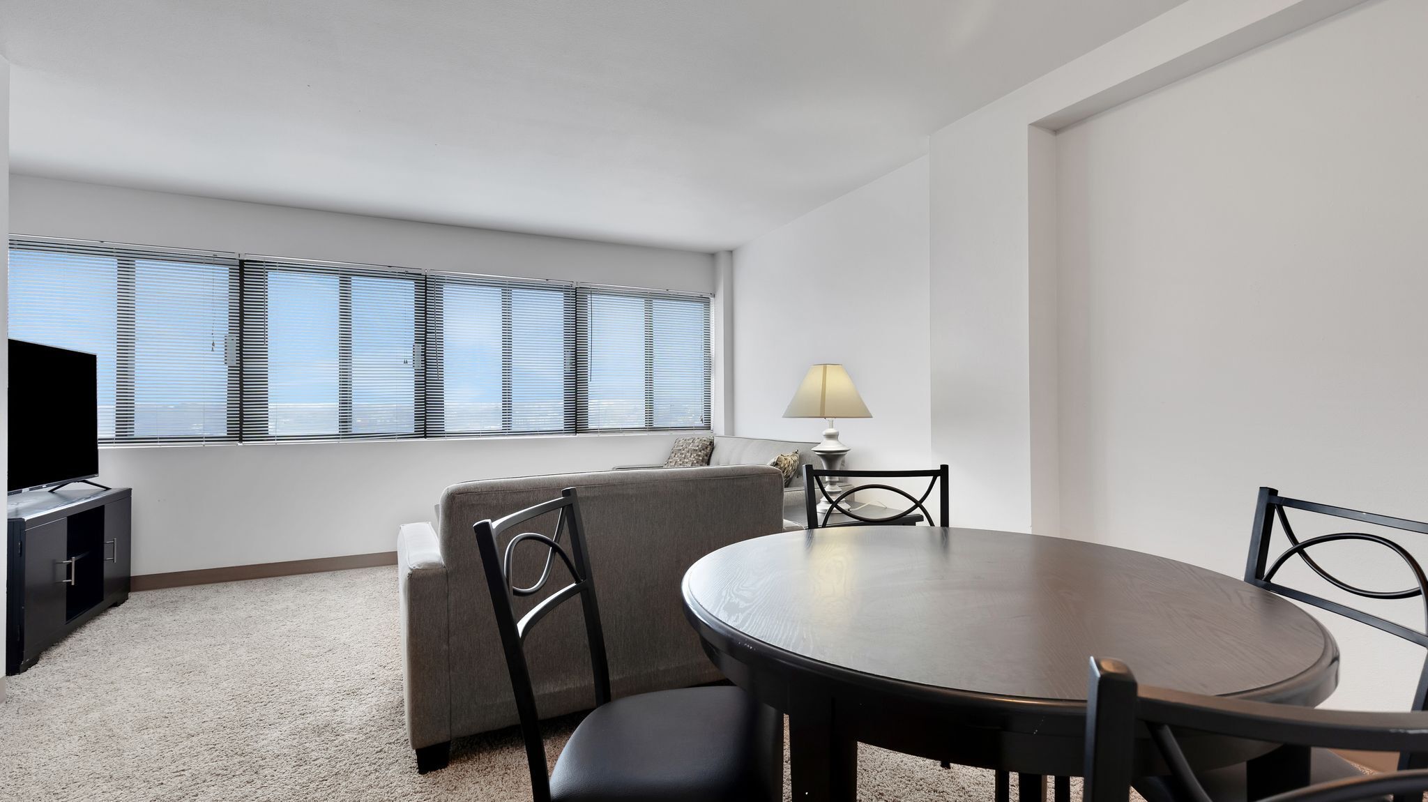 A modern hotel room with a round dining table, two chairs, a sofa, a TV, and a lamp.