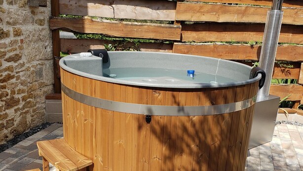 Un bain à remous en bois avec une structure métallique, un petit tabouret en bois et un mur de pierres en arrière-plan.