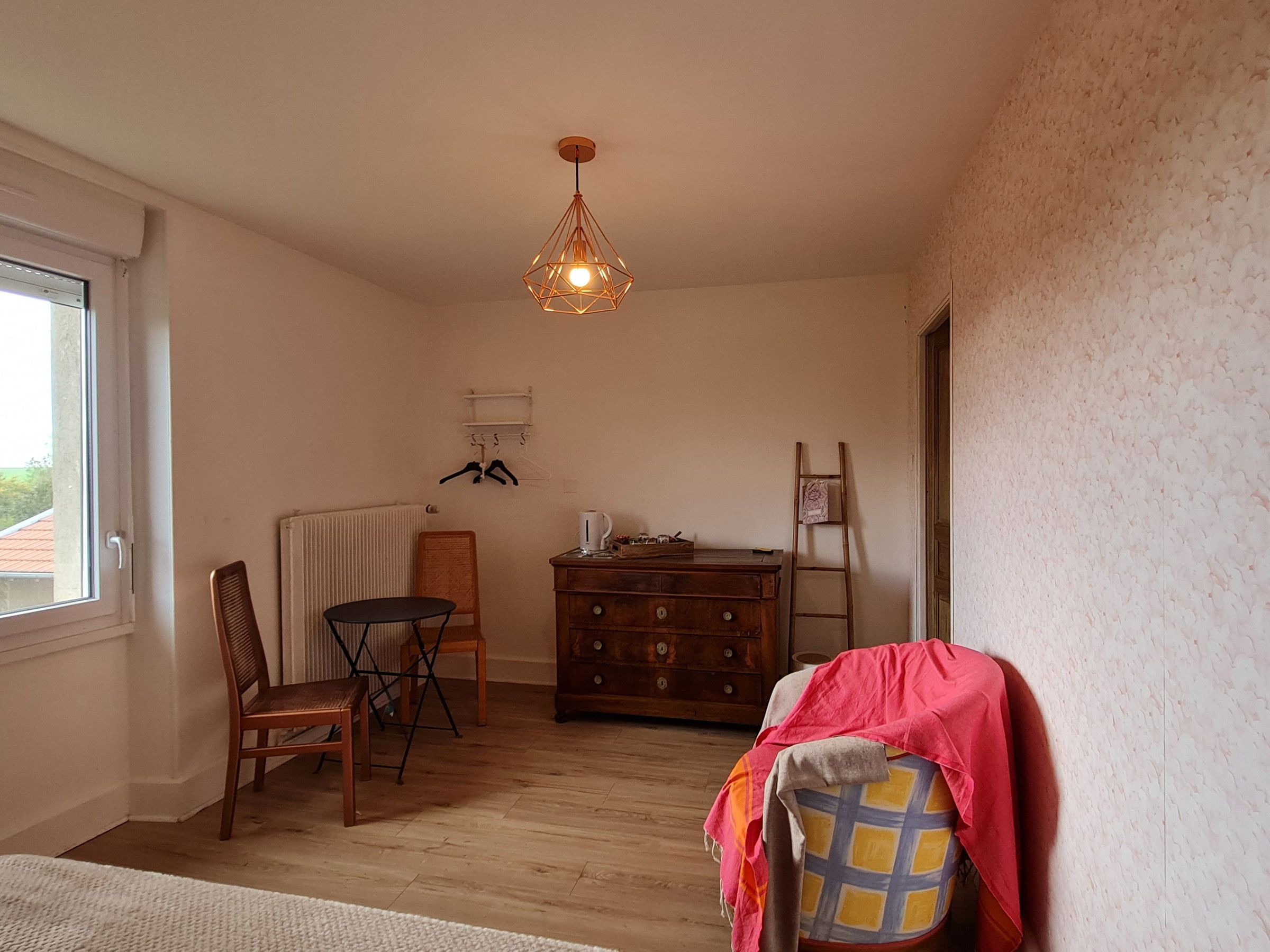 Een kamer met houten vloer, een radiator, een stoel, een tafel, een kast, een hanglamp en een raam.
