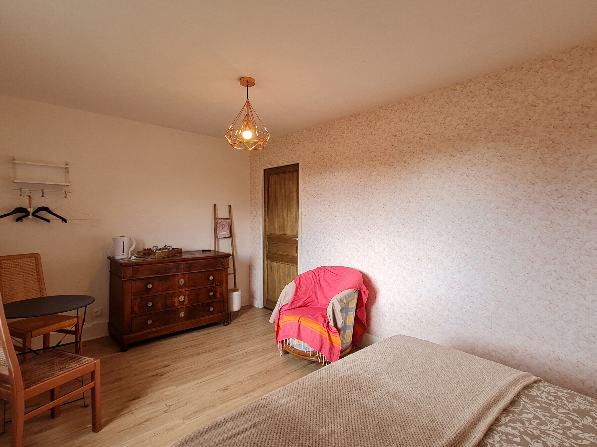 A bedroom with a wooden dresser, a chair, a bed with a patterned blanket, a hanging light fixture, and a door.