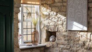 Una pared de piedra rústica con una ventana, un florero con plantas secas y una obra de arte abstracto enmarcada.