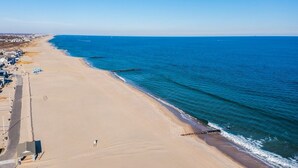 Een breed zandstrand met een heldere blauwe zee, enkele gebouwen langs de kust en een rotsachtige landtong in het water.