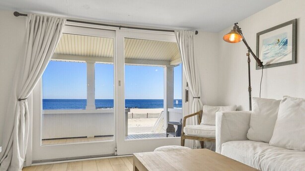 Een kamer met een grote glazen schuindeur die uitkomt op een uitzicht op het strand, een witte bank, een houten tafel en een lamp.