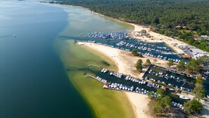 An aerial view of a coastal marina with boats docked, a sandy beach, and a forested area in the background. - Charming Pine Chalet - 4 Guests - Near Sanguinet Lake - Cleaning Included (Sanguinet)