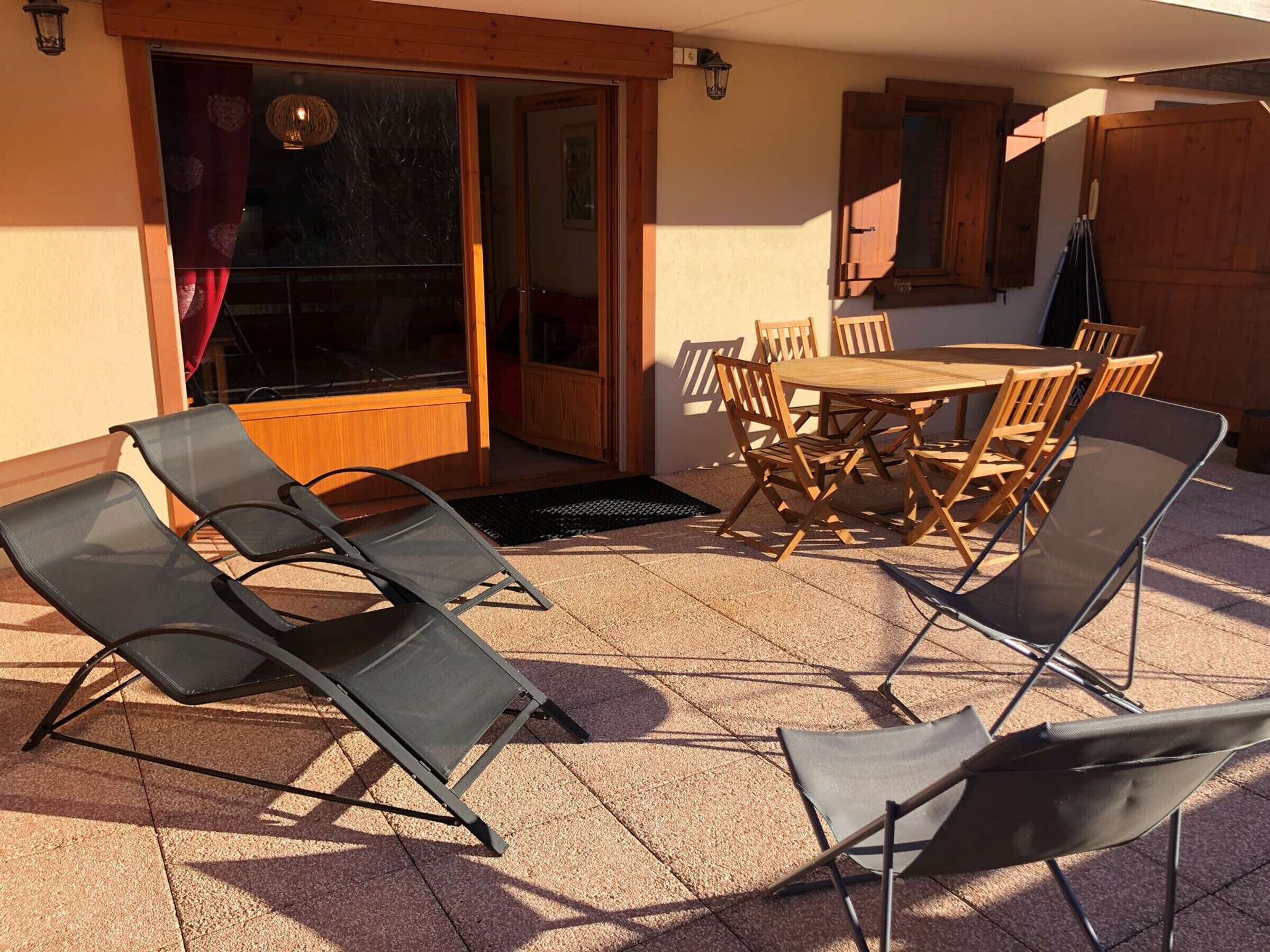 Een terras buiten met een houten tafel en stoelen, een schuifdeur en een klein houten kastje.