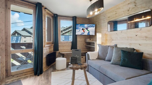 A cozy living room with a sofa, a coffee table, and a TV. Large windows offer a view of the outside. - Ski-in/Ski-out apartment with balcony and pets allowed (Val Thorens)