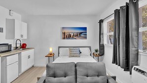 A modern bedroom with a grey sofa, a bed with white bedding, a nightstand with a lamp, a kitchen area with white cabinets, and a painting of a beach scene. - Young Circle & Hollywood Blvd ~ Posh Studio Suite (Hollywood)