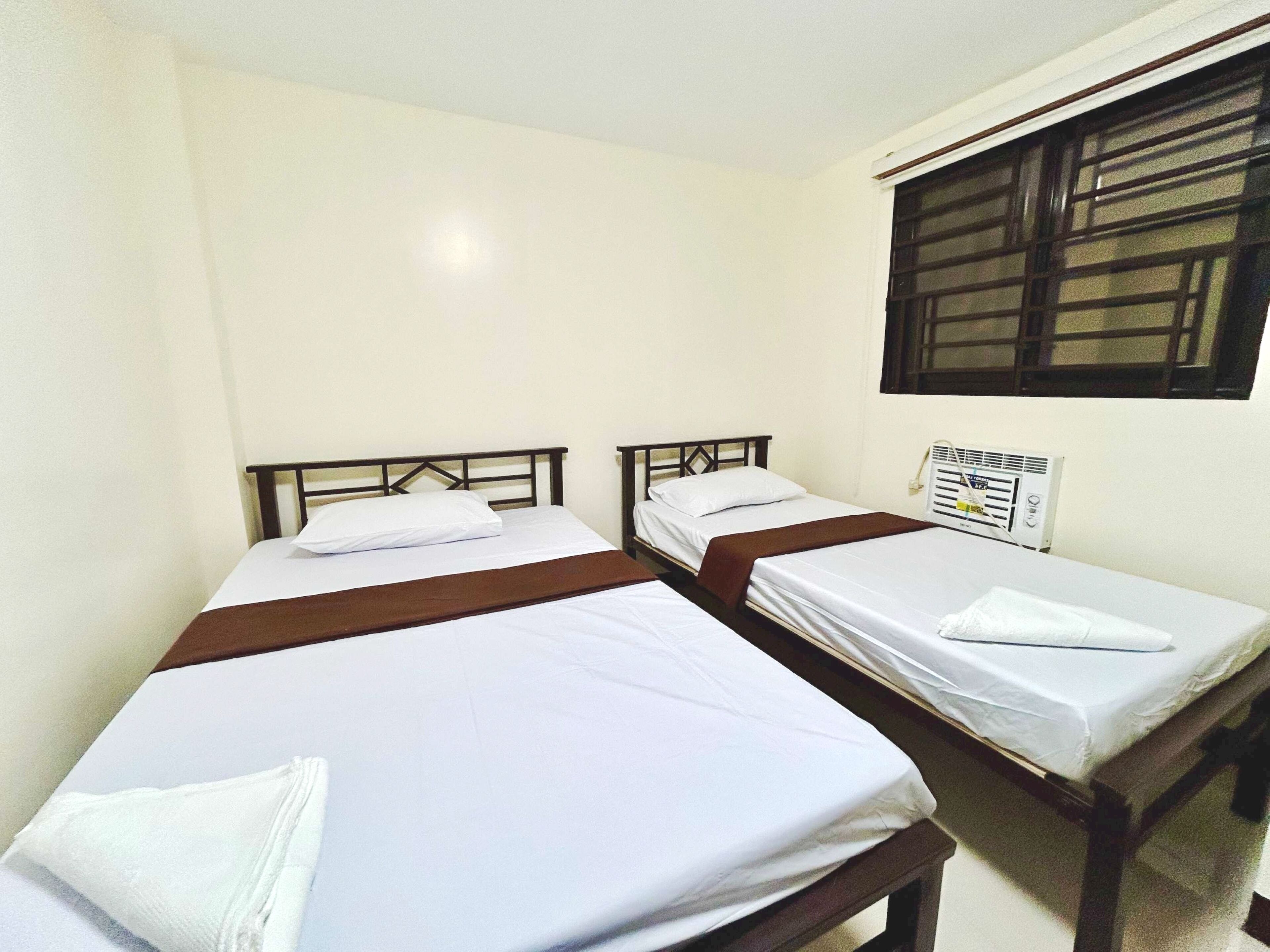 A room with two single beds, each with a wooden headboard and white bedding. There is a window with blinds on the wall, and a white radiator is visible.