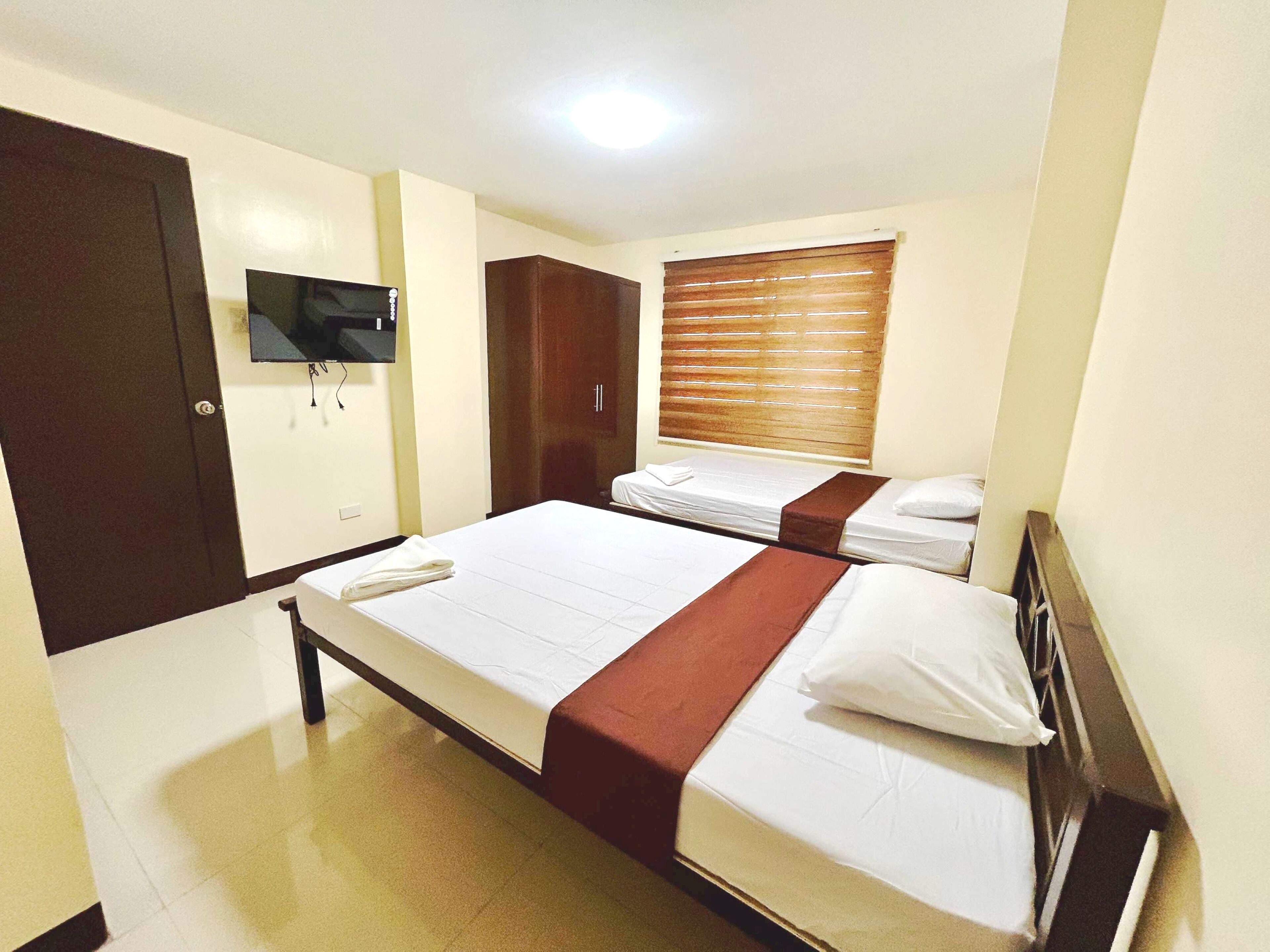 Habitación de hotel con dos camas, un televisor montado en la pared, un armario y una ventana con persianas.