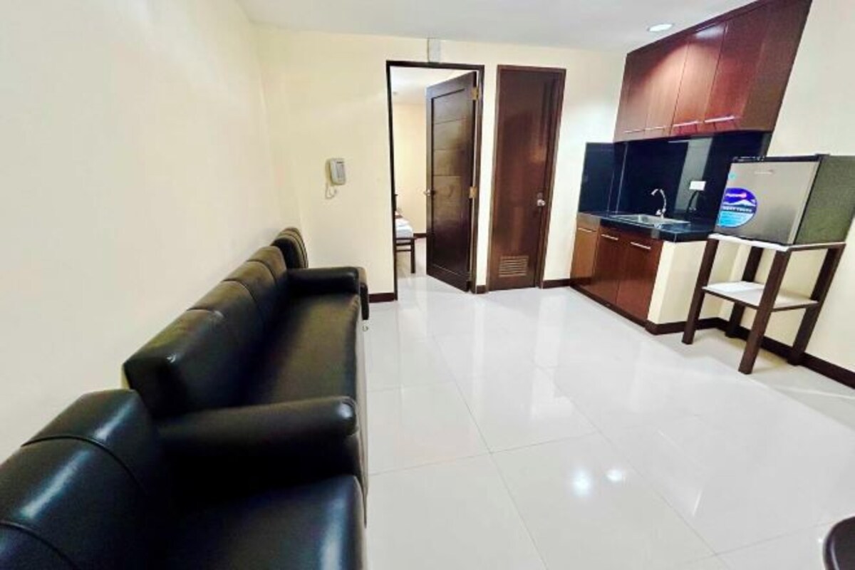 A modern living room with a black leather sofa, a kitchen area with a refrigerator, and a door leading to another room.