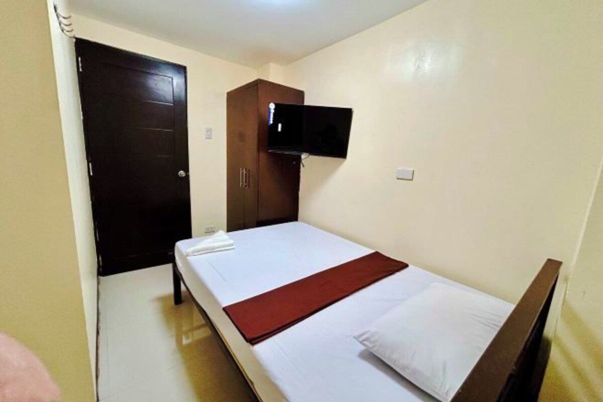 A single bed with a white mattress and a red blanket, a dark wooden wardrobe, and a flat-screen TV mounted on the wall.