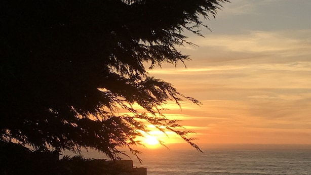 Une vue sur le coucher de soleil depuis un balcon donnant sur l’océan, avec une balustrade en bois et de la végétation au premier plan.