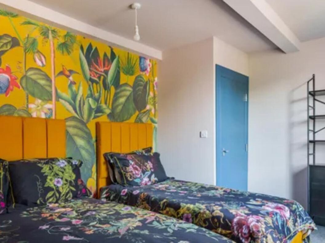 A room with a bed featuring floral bedding, a vibrant tropical mural on the wall, and a blue door.