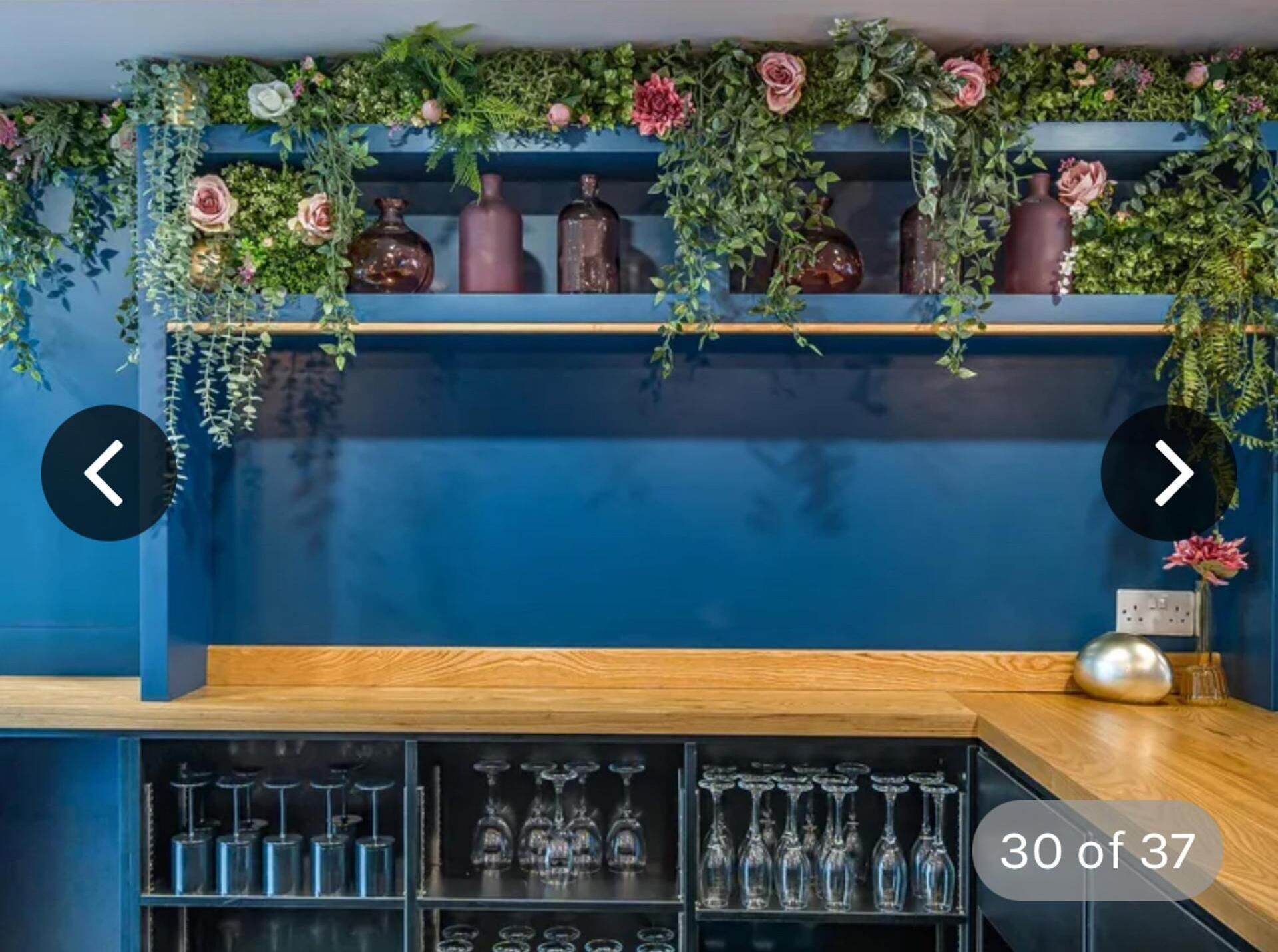 A bar area with shelves of glassware and decorative vases, a wooden counter, and a blue wall.