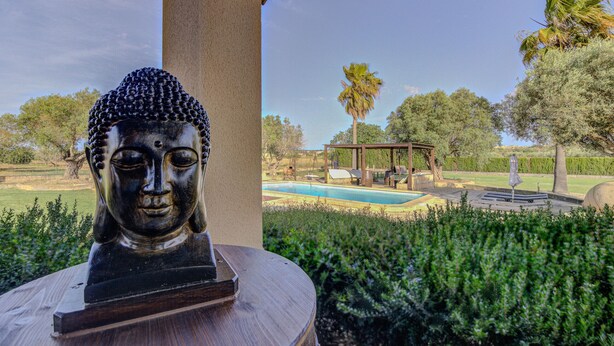 Une statue de Bouddha sur un piédestal en bois, dans un jardin avec une piscine et un kiosque en arrière-plan.