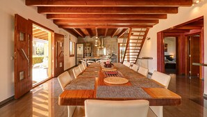 Un vaste espace dédié à la salle à manger, avec une longue table en bois, des chaises blanches et un plafond haut avec des poutres apparentes.