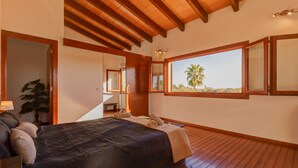 Une chambre avec un grand lit, un plancher en bois, une vue sur des palmiers et un plafond en bois.