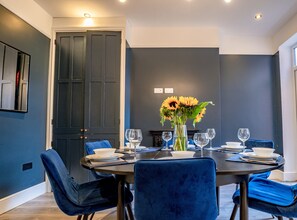 A dining area with a dark-colored table set for four, blue chairs, and a vase with sunflowers.