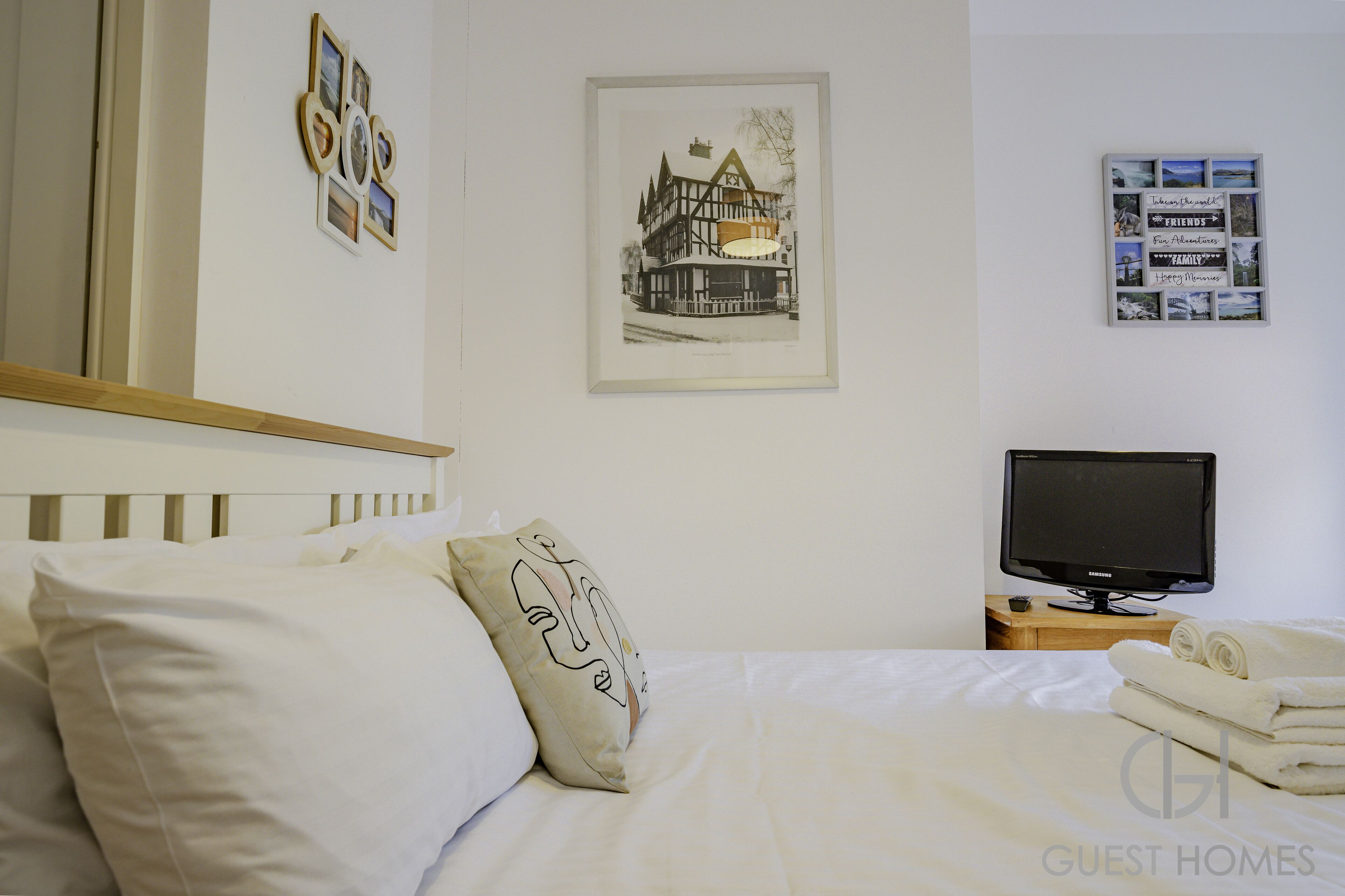 A neatly made bed with white linens, a television on a wooden stand, and framed pictures on the wall.