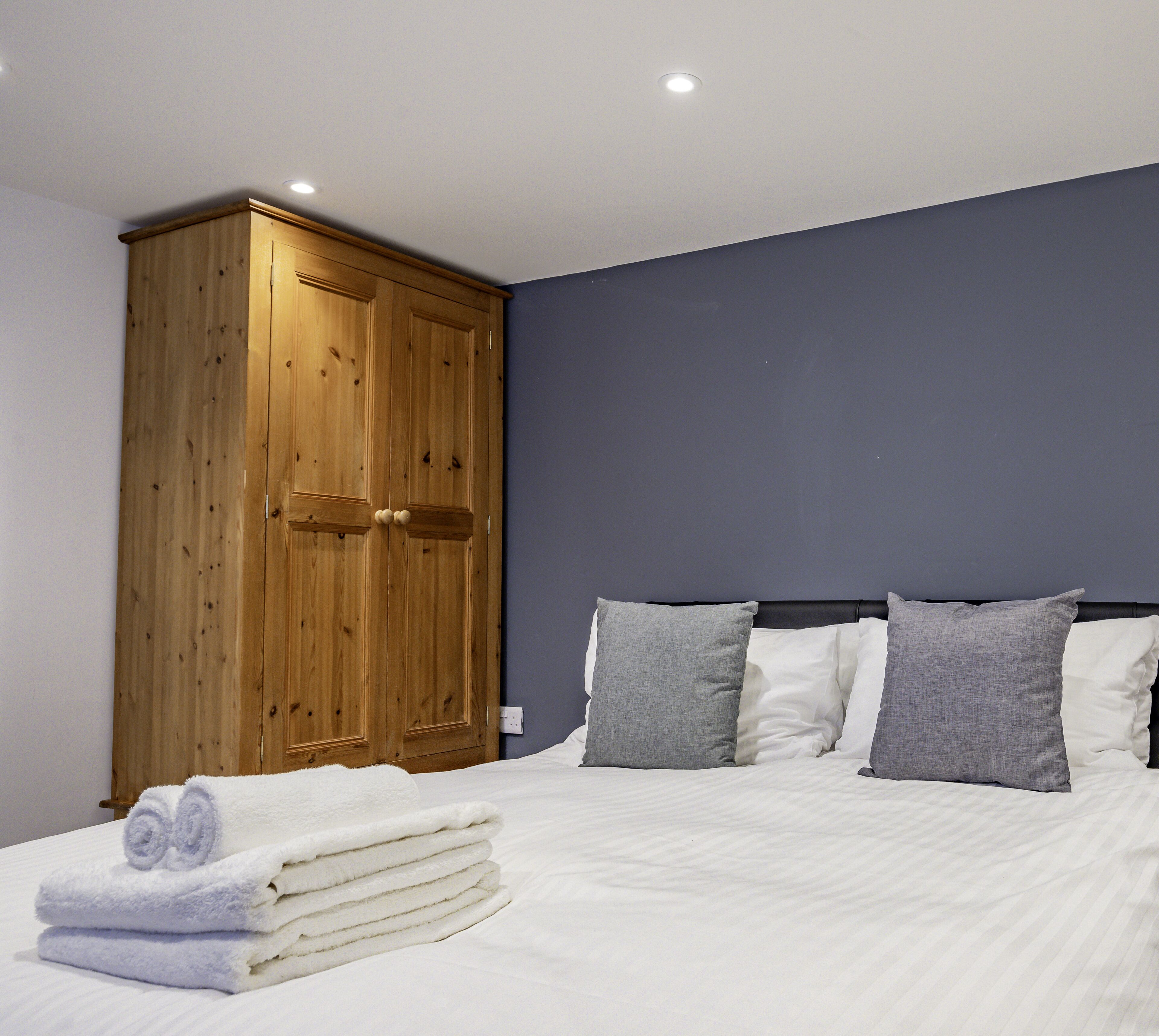 Un dormitorio con una cama, almohadas grises, un armario de madera y toallas enrolladas.