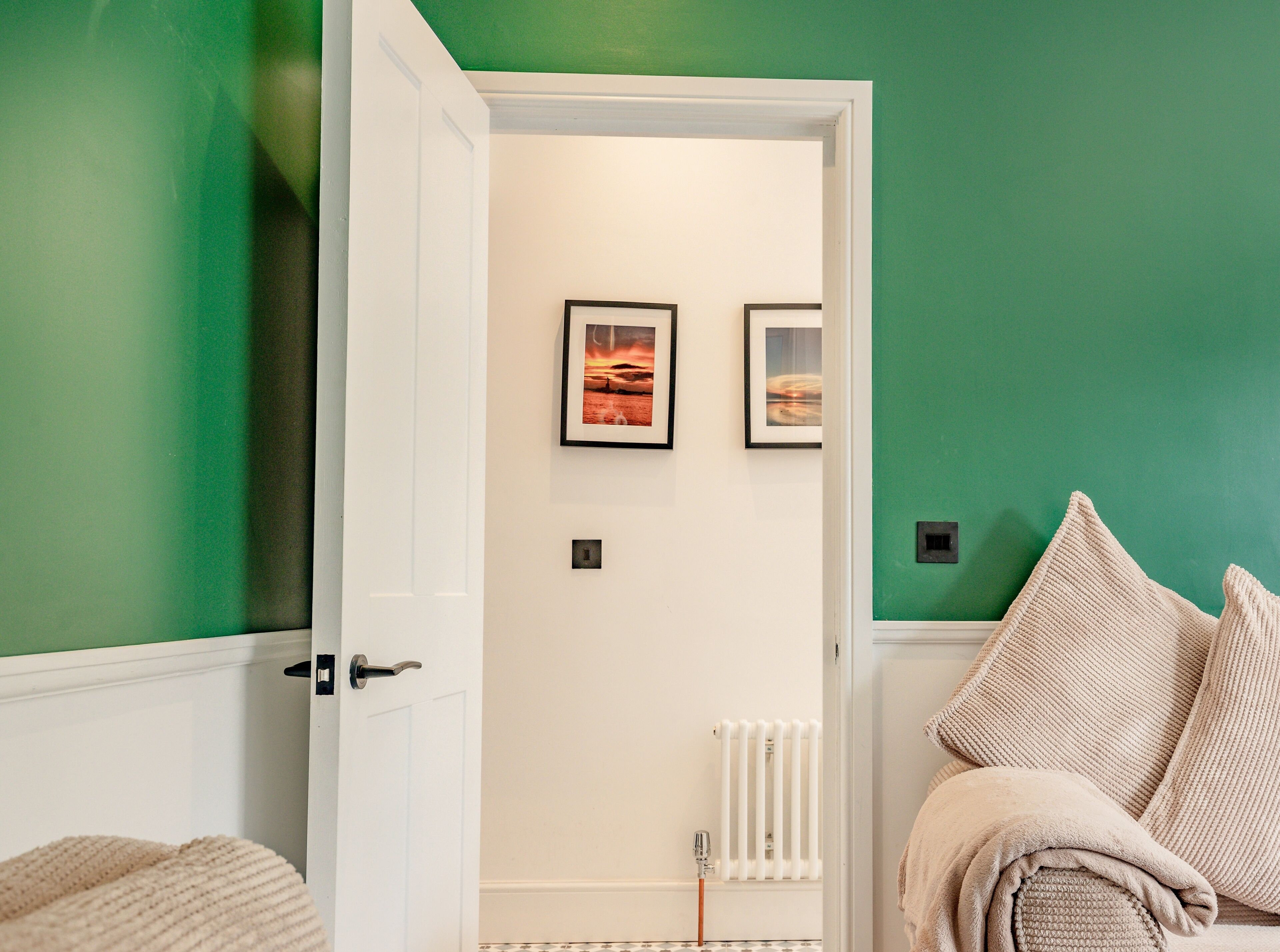 A room with a green wall, white door, and a radiator. There are two framed pictures on the wall and a beige blanket on the floor.