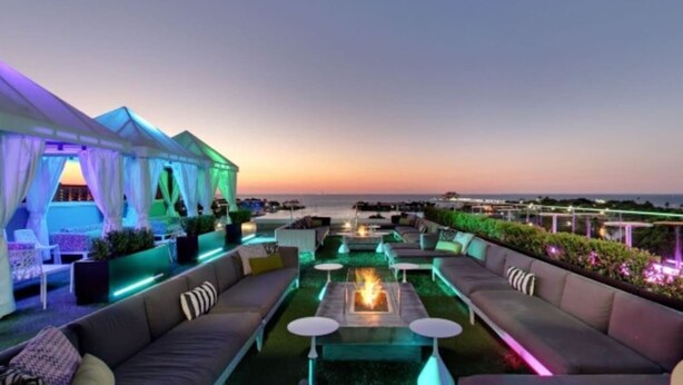 A rooftop terrace with outdoor seating, a fire pit, and a view of the ocean at sunset.