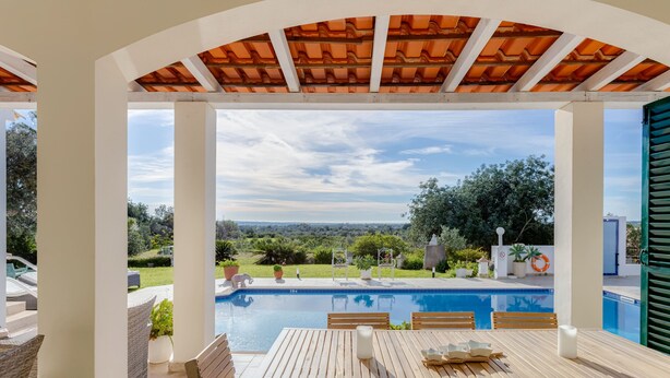 Área de piscina ao ar livre com mesa e cadeiras de madeira, cercada por pilares brancos e uma pérgula com telhado de azulejos.
