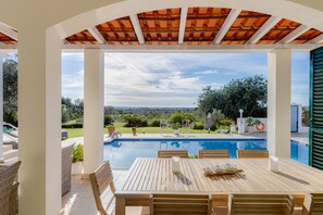 Ein Außenpoolbereich mit Holztisch und -stühlen, umgeben von weißen Säulen und einer Pergola mit Ziegeldach.