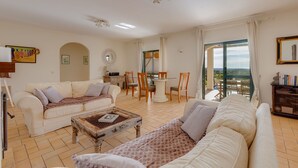 Uma sala de estar ampla, com um sofá branco, uma mesa de centro e uma área de jantar com mesa e cadeiras.