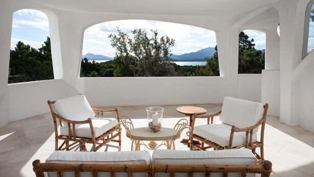 A room with white walls, a round table, and four wicker chairs with white cushions. There are large windows offering a view of trees and mountains. - MANOR HOUSE (ARZACHENA LOC. CAPRICCIOLI)
