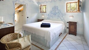 A bedroom with a large bed, a bedside table, a lamp, a small cabinet, a wicker chair, and a window with a view of trees. - MANOR HOUSE (ARZACHENA LOC. CAPRICCIOLI)
