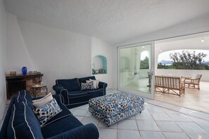Un espacio de estar con un sofá, un reposapiés y un ventanal de vidrio que da a un patio exterior con una mesa y sillas.