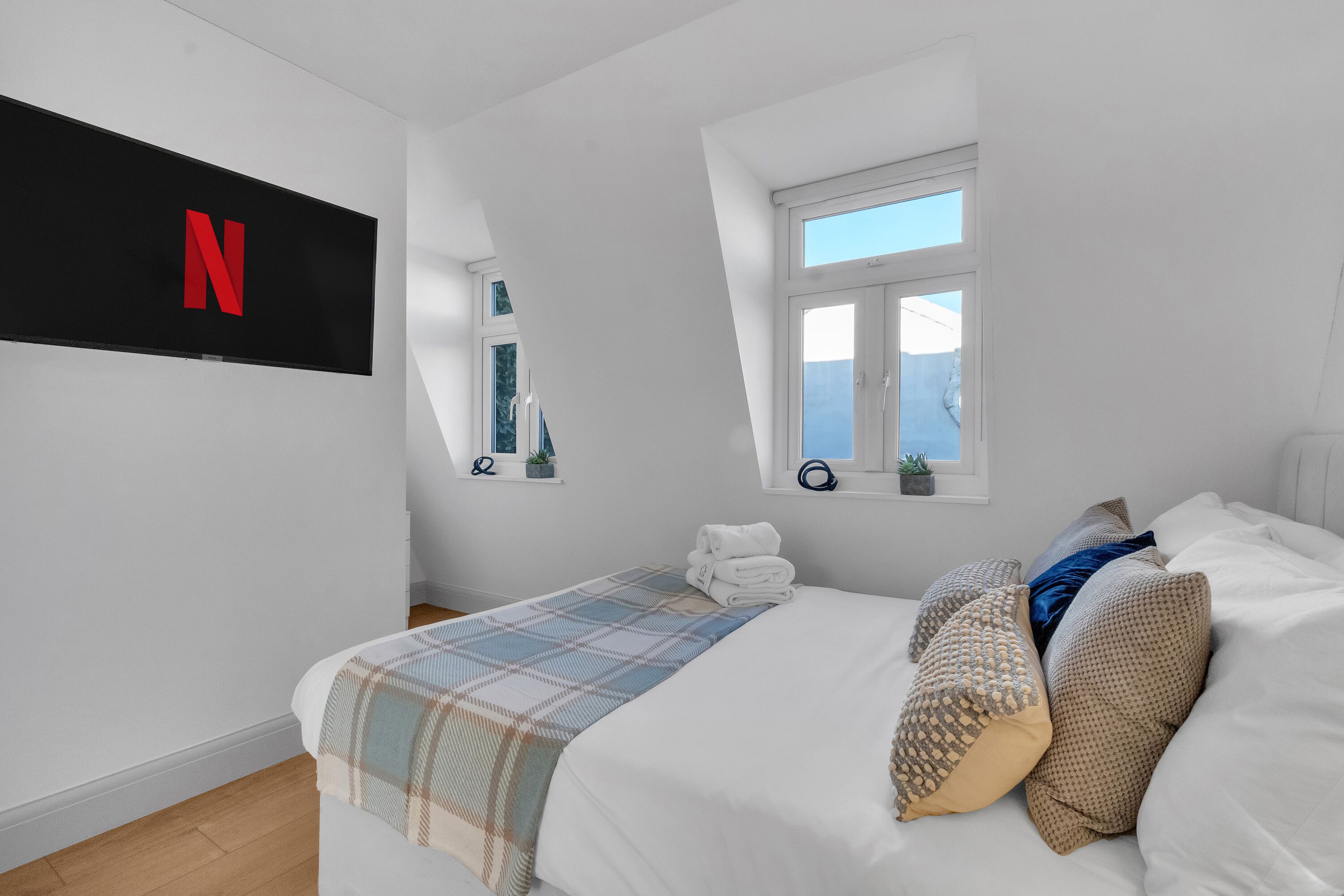 Un dormitorio moderno con una cama grande, un televisor montado en la pared, un ventanal pequeño y una estantería con objetos decorativos.