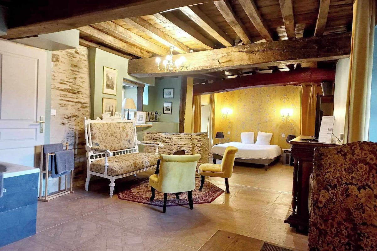 Quarto com cama, duas poltronas, um sofá, teto de madeira e parede de pedra.
