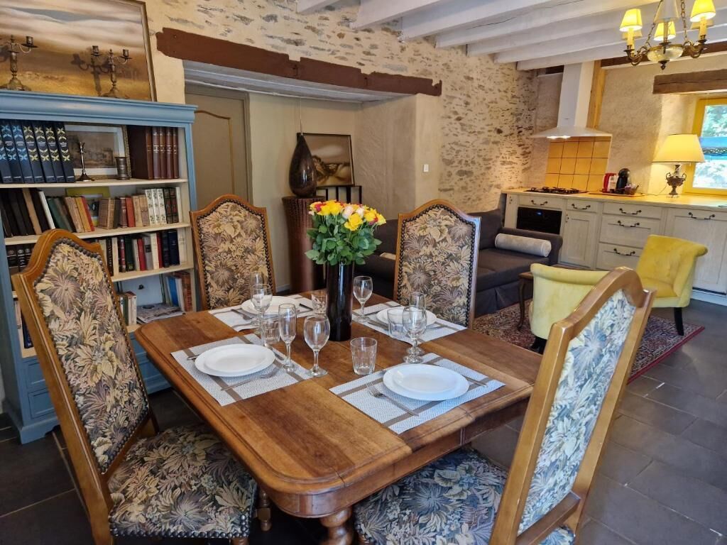 A dining area with a wooden table set for four, surrounded by floral-patterned chairs. A bookshelf with books and decorative items is to the left.
