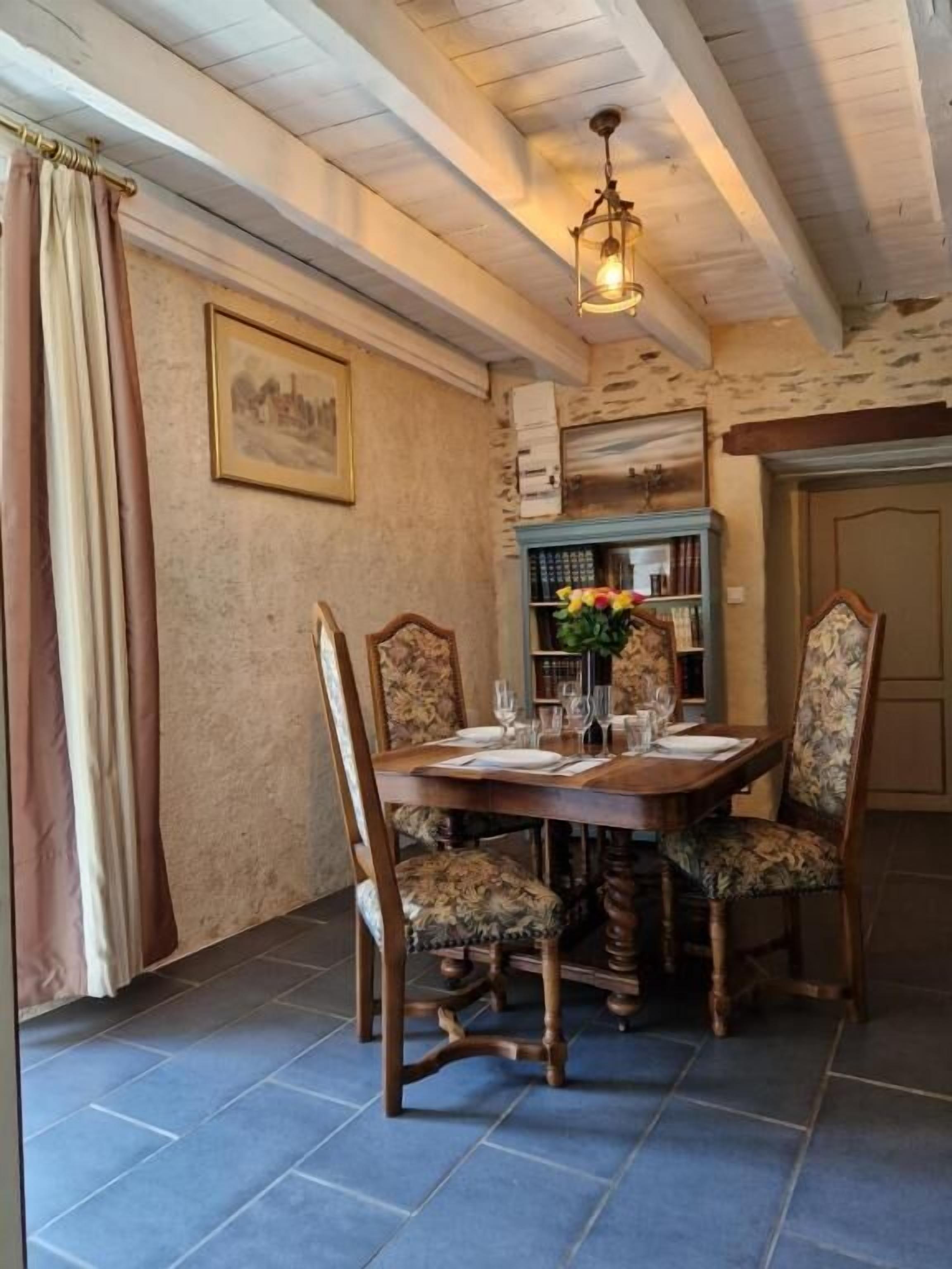 A dining area with a wooden table set for four, surrounded by upholstered chairs. A chandelier hangs from the ceiling, and a bookshelf with books and a vase of flowers is visible in the background.